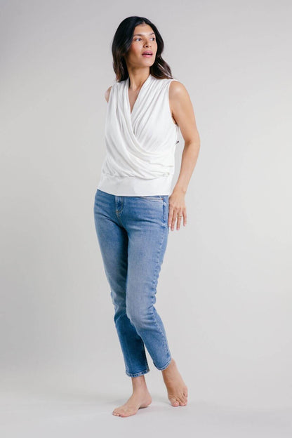 A woman modeling a white draped sleeveless top and light blue cropped jeans against a plain light gray studio background.