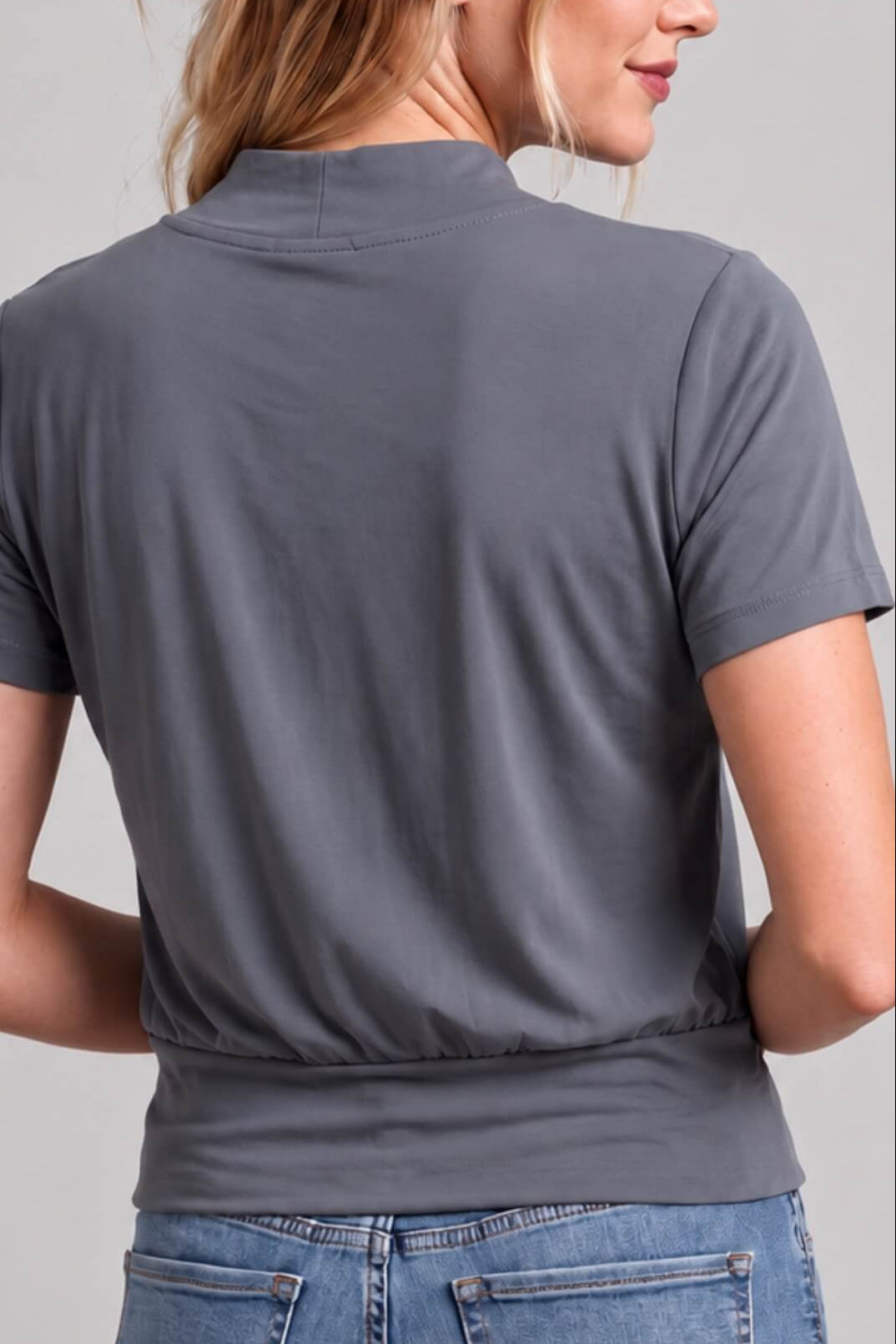A woman shown from the back wearing a dark gray short-sleeve mock neck top and blue jeans against a light gray studio background.