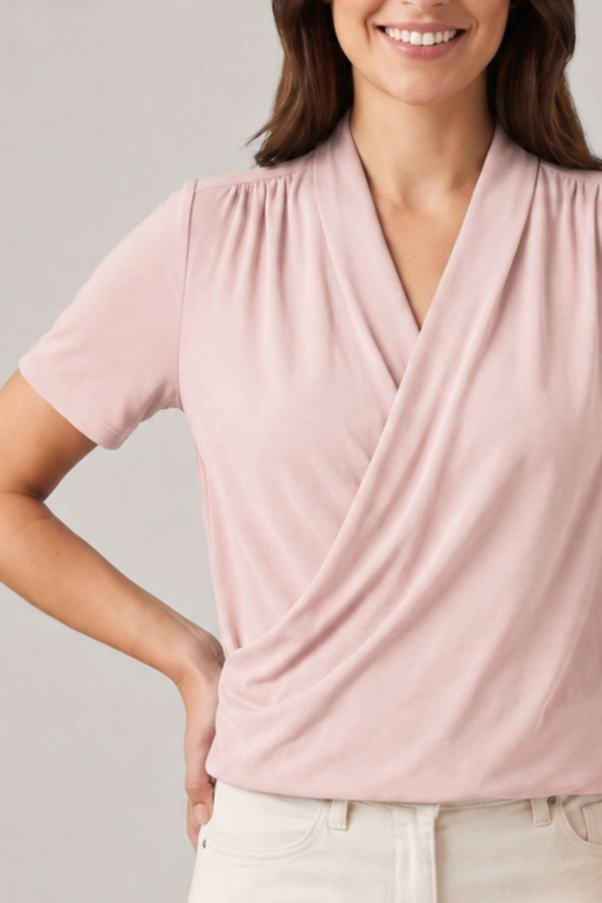 A woman wearing a short-sleeve wrap-style blouse in a soft dusty pink color, paired with light beige pants, against a neutral gray background.