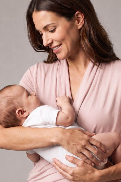 A woman in a dusty pink wrap top holds a baby wearing a white onesie against a neutral gray background.
