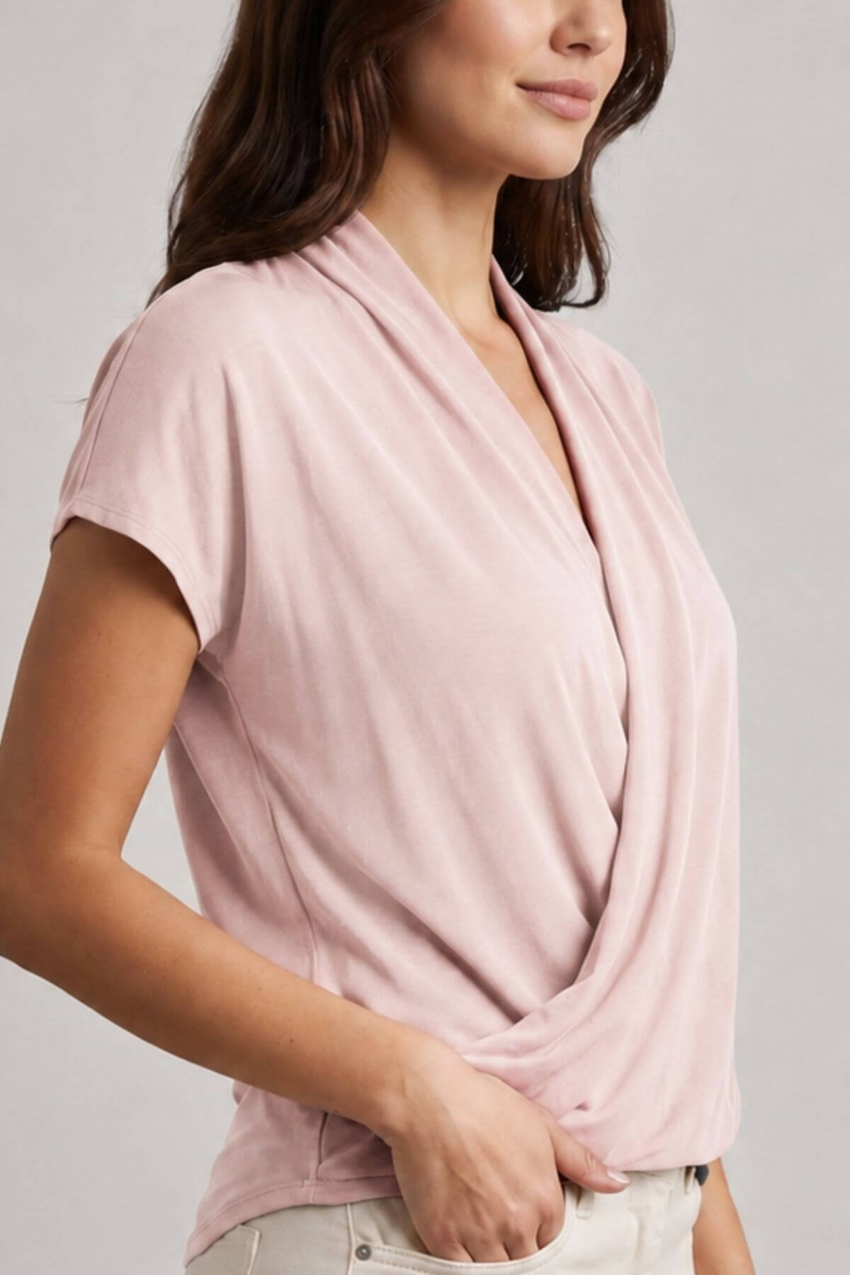 A woman wearing a light pink short-sleeve wrap-style top with a draped neckline, paired with light-colored pants, photographed against a plain light gray background.