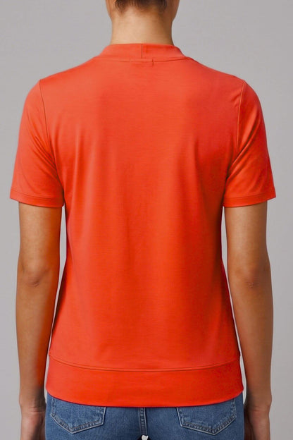 A person shown from the back wearing a bright orange short-sleeve polo shirt and blue jeans against a plain gray studio background.