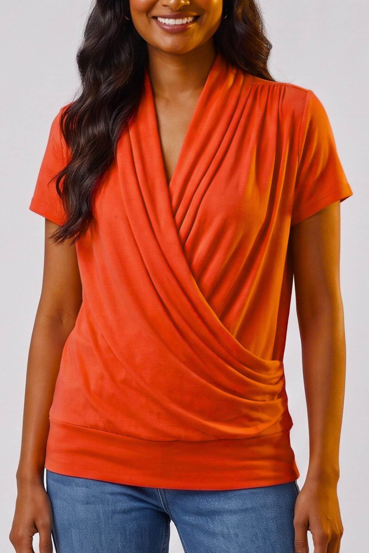 A woman wearing a bright orange short-sleeve wrap-style top with a draped front, paired with blue jeans, against a light gray studio background.