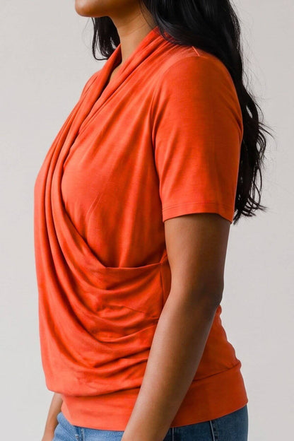 A woman wearing a short-sleeve orange top with a draped, wrap-style front and side pockets, paired with blue jeans against a plain light gray background.