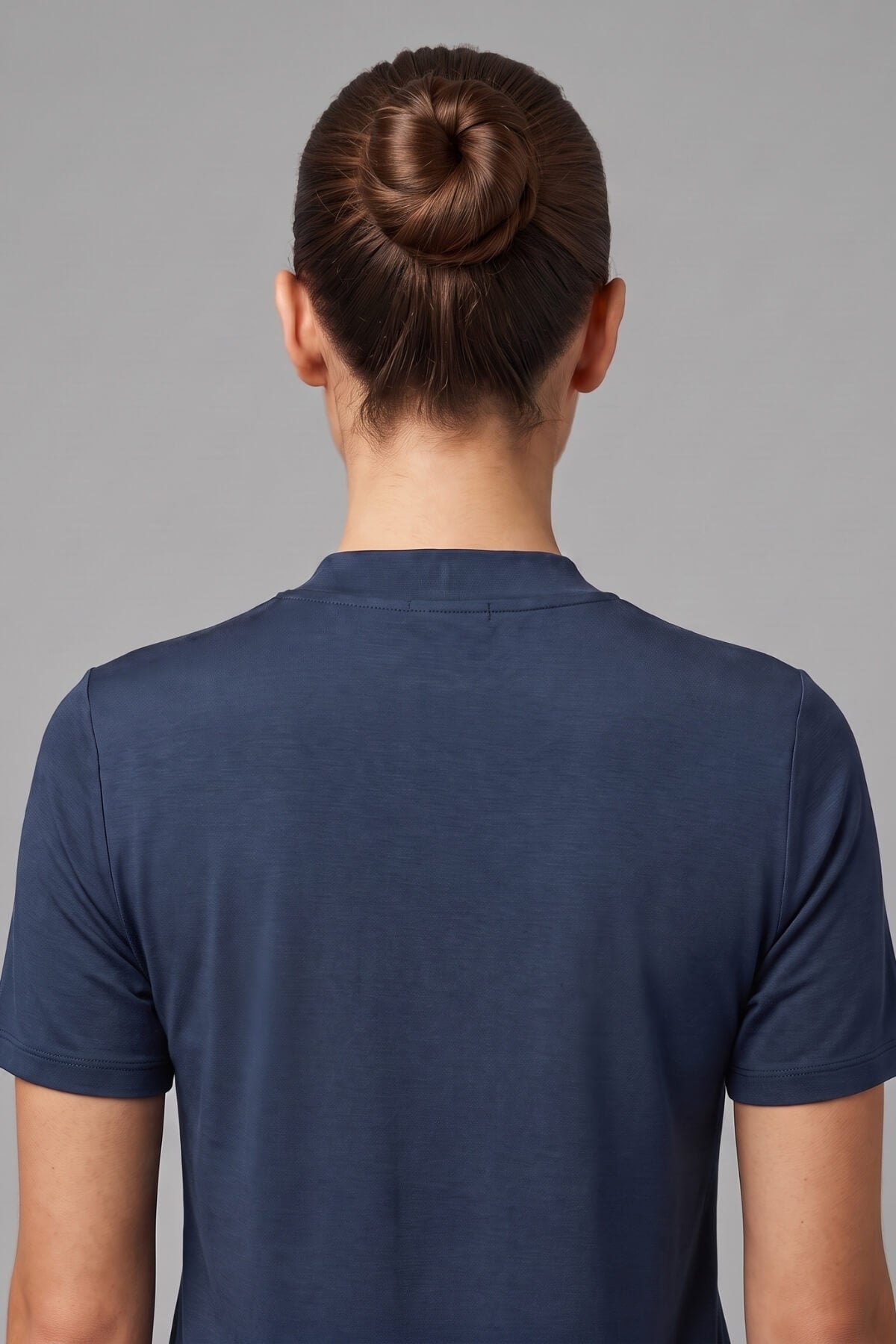 Back view of a person wearing a navy blue short-sleeve crewneck t-shirt, with dark brown hair styled in a bun, against a solid gray studio background.