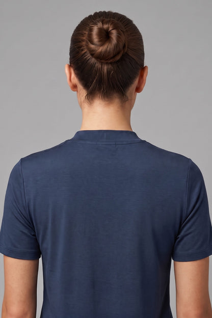 Back view of a person wearing a navy blue short-sleeve crewneck t-shirt, with dark brown hair styled in a bun, against a solid gray studio background.