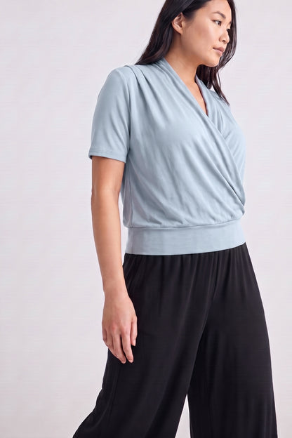 A woman wearing a light blue short-sleeve wrap-style top and black wide-leg pants, photographed against a plain light gray studio background.