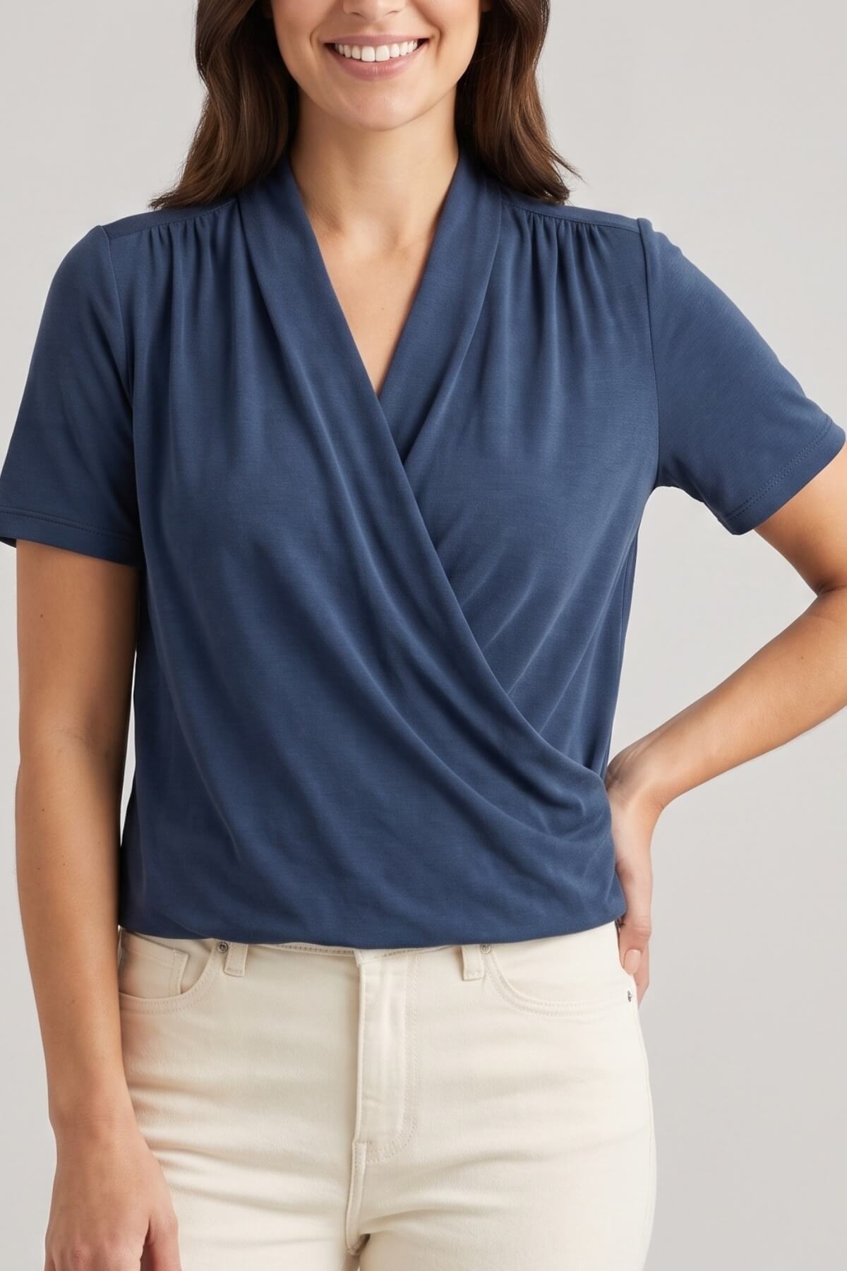 A woman wearing a navy blue short-sleeve wrap-style blouse with a V-neck and gathered neckline, paired with light beige pants, against a plain light gray studio background.