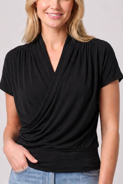 A woman wearing a black short-sleeve wrap-style top with a draped V-neckline, paired with light blue jeans, against a plain light gray background.