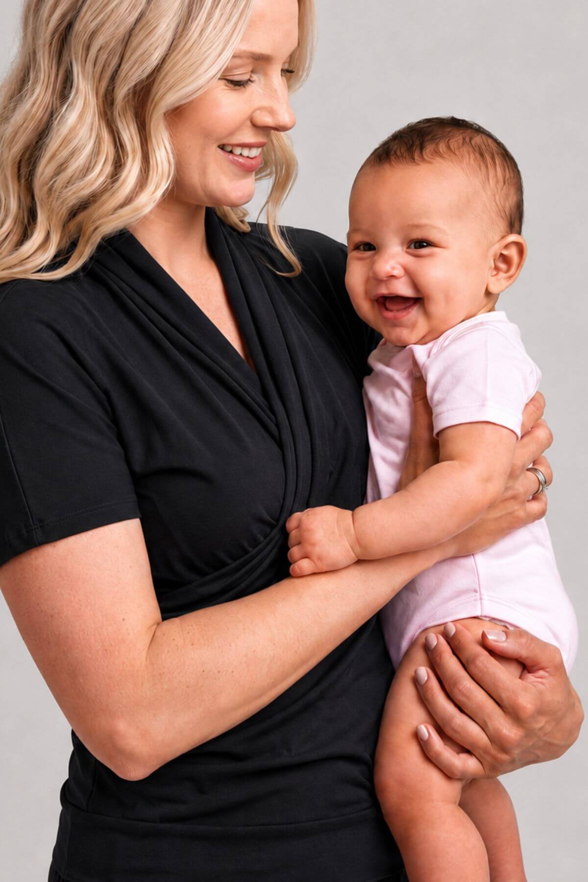 A smiling blonde woman in a black wrap-style top holds a baby wearing a light pink short-sleeve bodysuit against a plain light gray studio background.