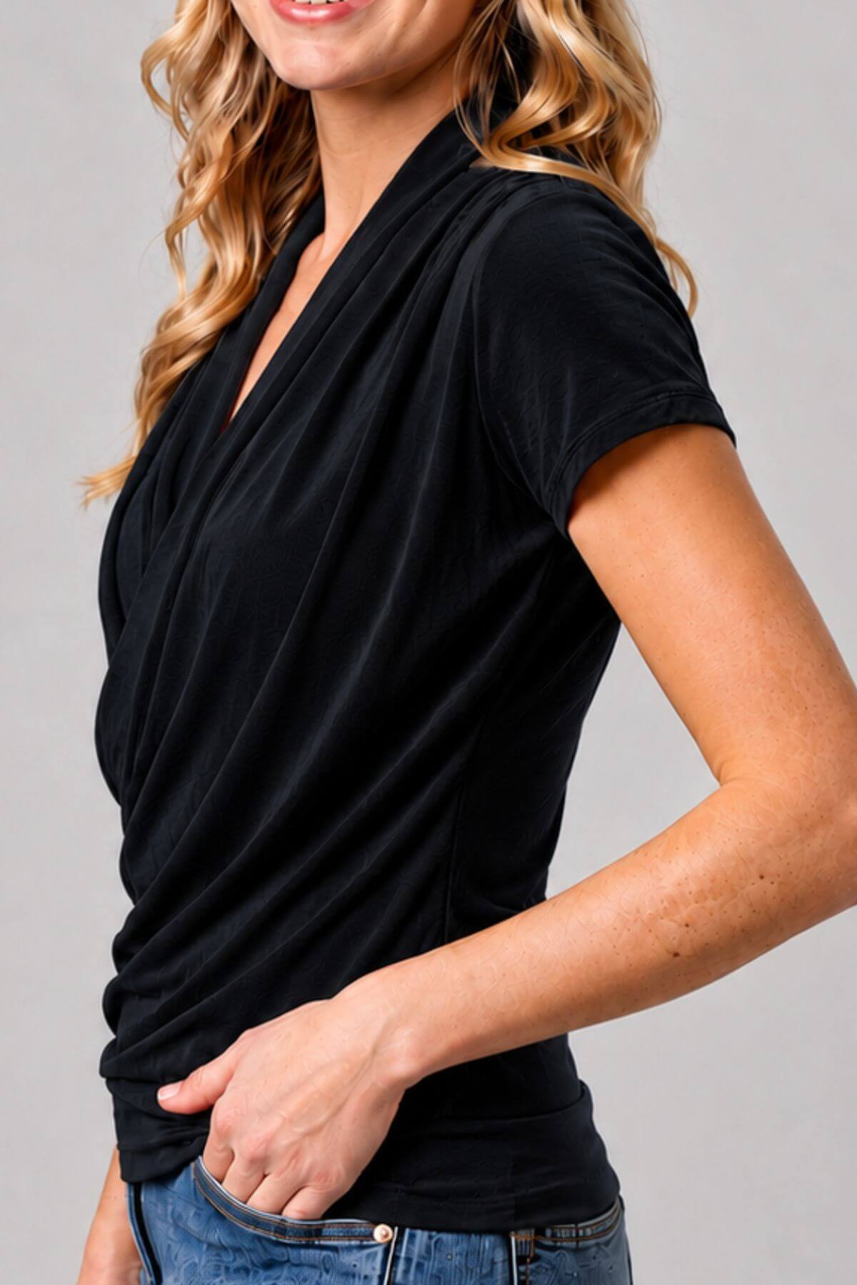A woman wearing a black short-sleeve wrap-style top with a draped neckline, paired with blue jeans, shown against a plain light gray studio background.