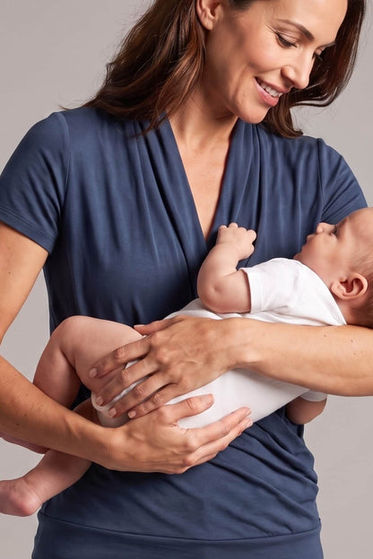A woman wearing a navy blue wrap-style baby shirt holds a baby dressed in a white onesie against a plain light gray background.