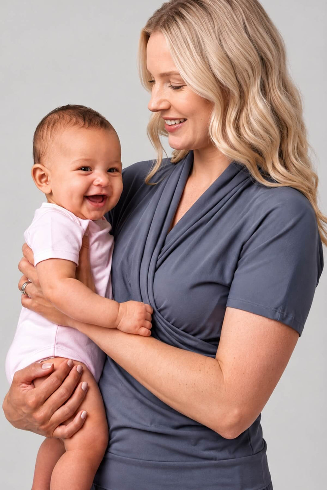 A woman holding a smiling baby, wearing a slate blue-gray short-sleeve wrap-style top. The baby is dressed in a light pink onesie.