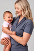 A woman holding a smiling baby, wearing a slate blue-gray short-sleeve wrap-style top. The baby is dressed in a light pink onesie.
