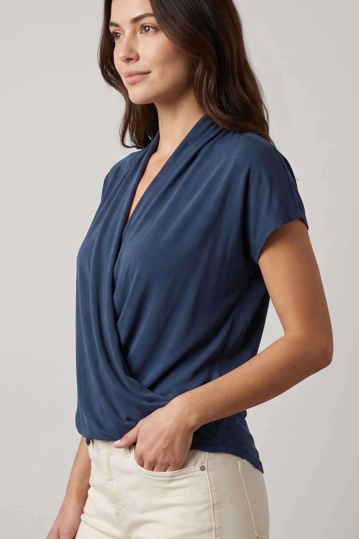 A woman wearing a navy blue short-sleeve wrap-style blouse and cream pants, photographed against a plain light gray studio background.