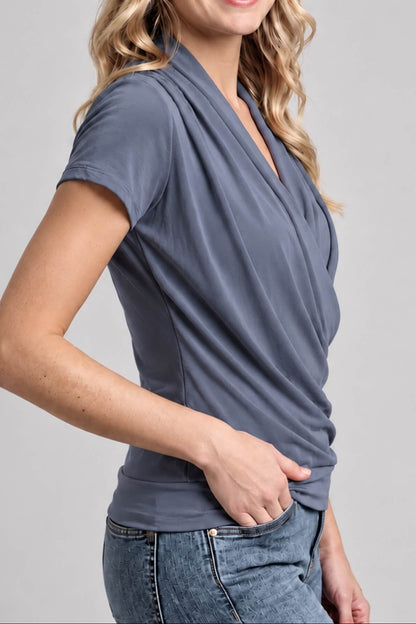A woman wearing a slate blue-gray short-sleeve wrap-style V-neck top and blue jeans, shown from the side against a light gray studio background.