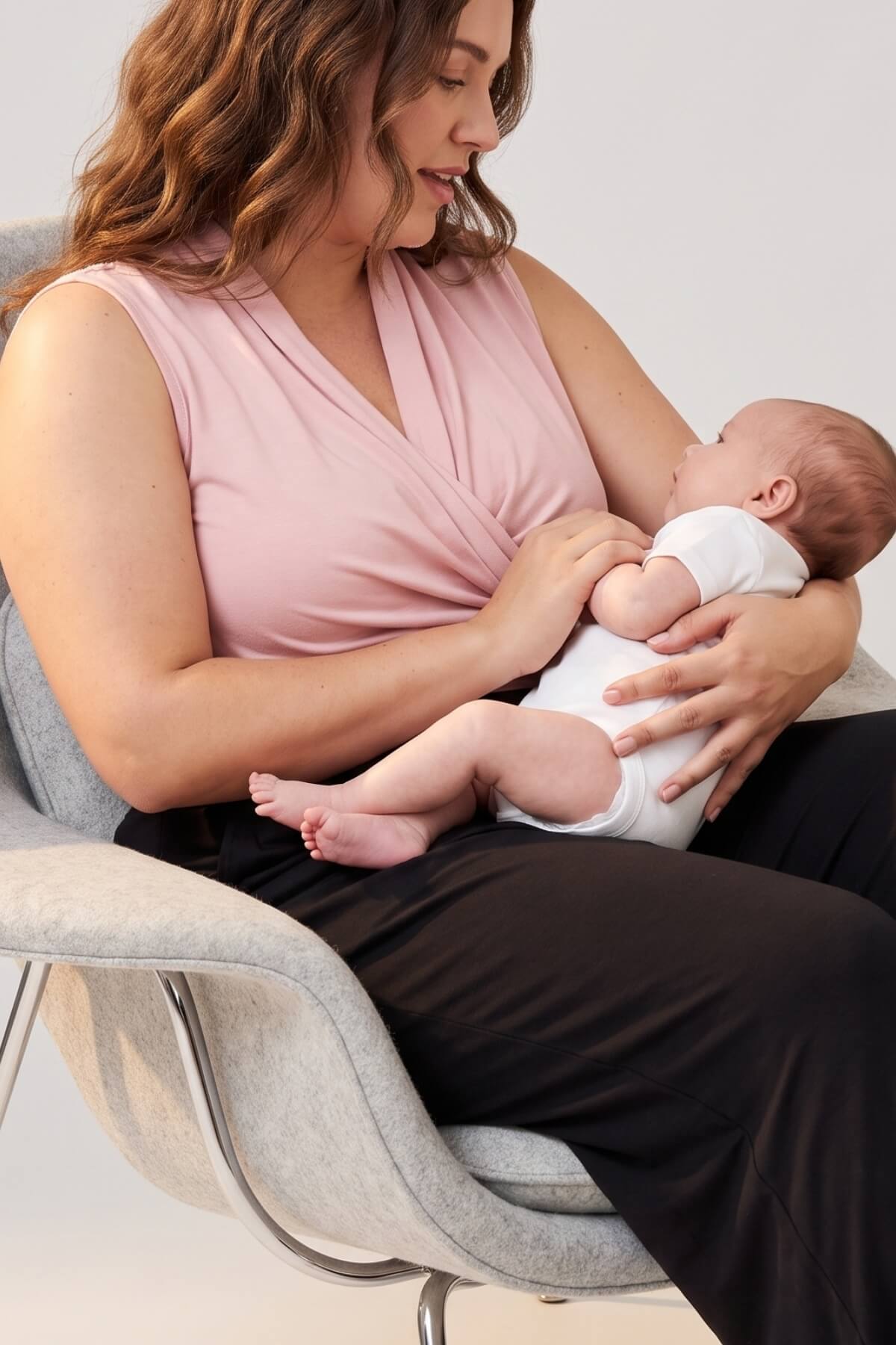 A woman in a light pink sleeveless top and black pants sits in a gray chair, breastfeeding a baby wearing a white onesie.