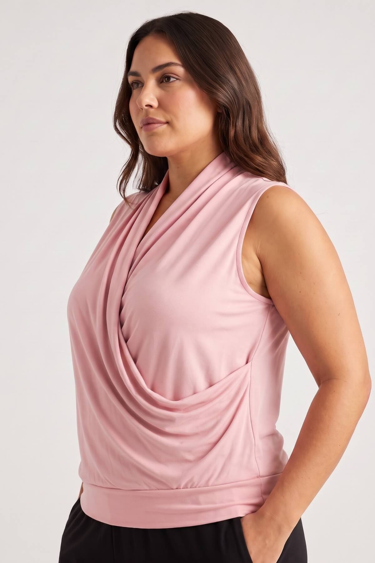 A woman wearing a sleeveless dusty pink wrap-style top with a draped front, shown from the waist up against a plain light gray studio background.