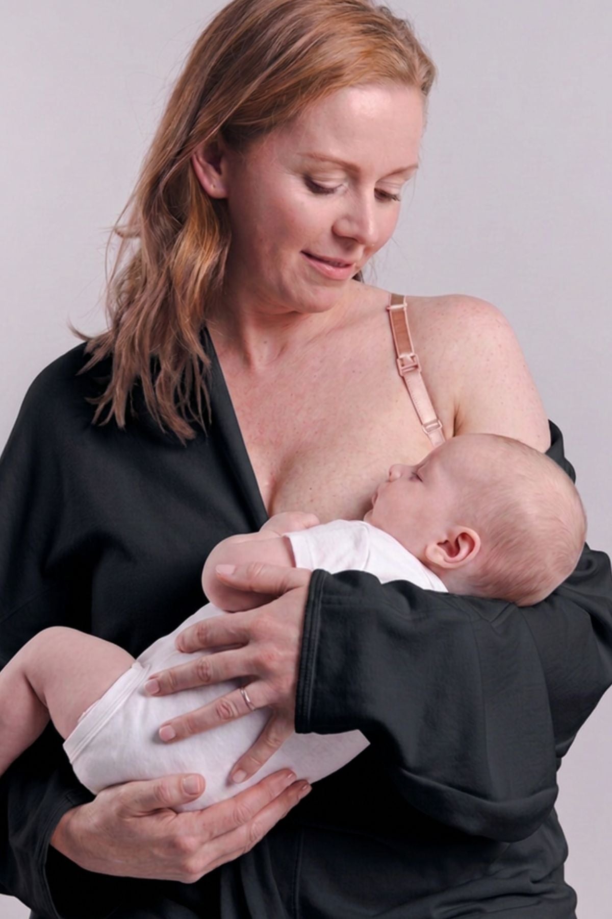 A woman wearing a black nursing top holds a baby wearing a white outfit against a plain light gray background.