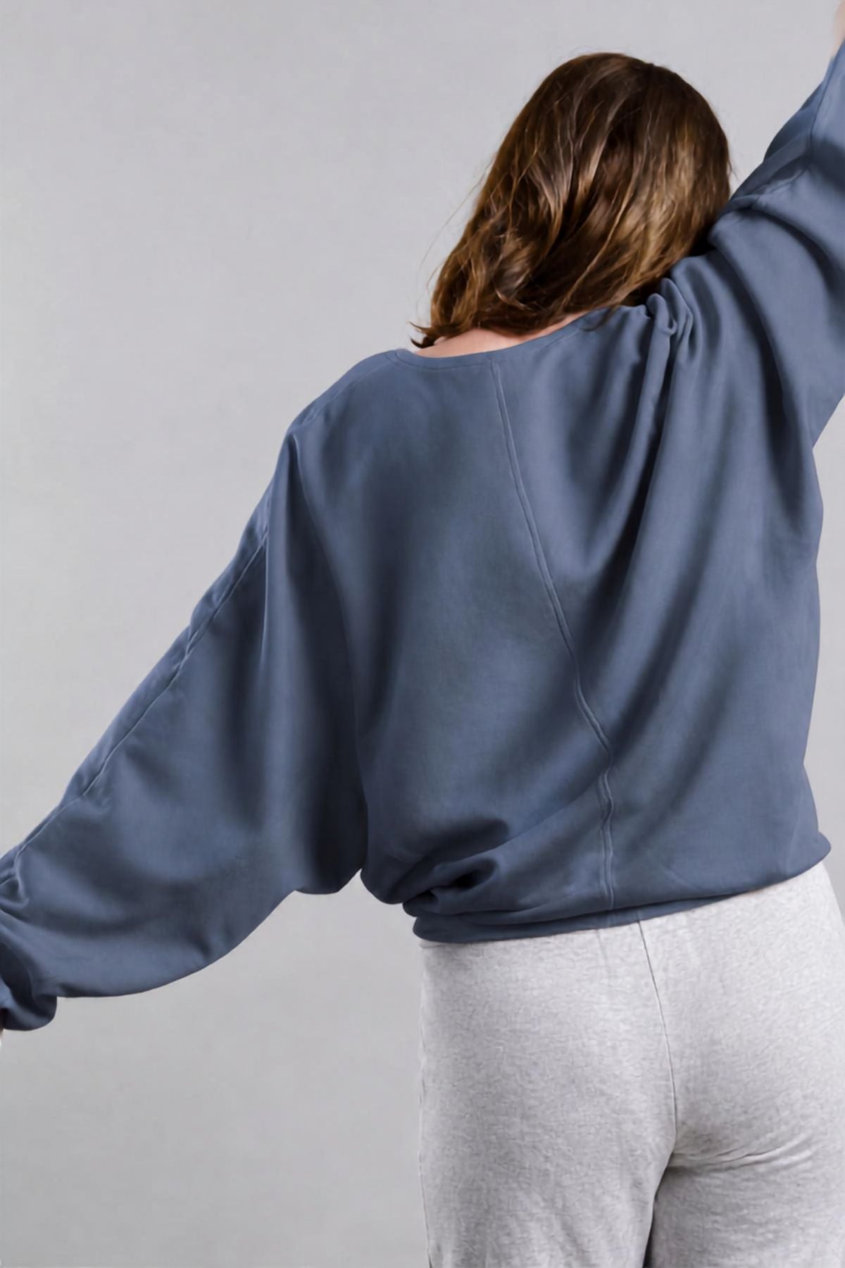 A woman shown from behind wearing a slate blue cropped sweatshirt and light gray sweatpants against a neutral gray background.