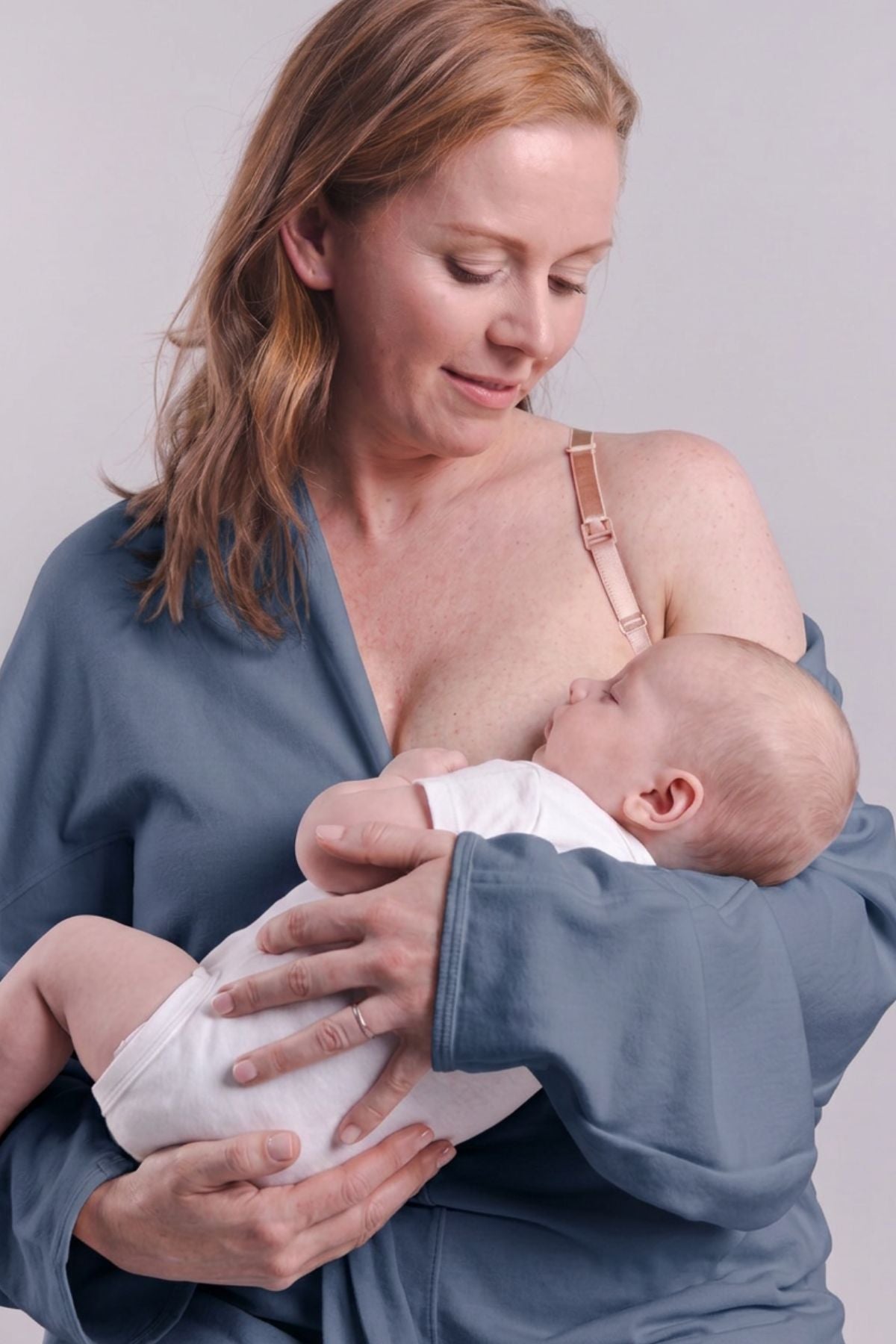 A woman in a slate blue nursing robe holds a baby wearing a white bodysuit while breastfeeding. The woman has red hair and is wearing a light pink bra underneath the robe.