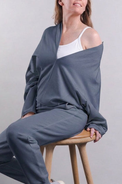 A woman sits on a wooden stool wearing a matching gray lounge set consisting of a draped long-sleeve top and jogger pants over a white tank top and white sneakers, against a plain light gray wall.