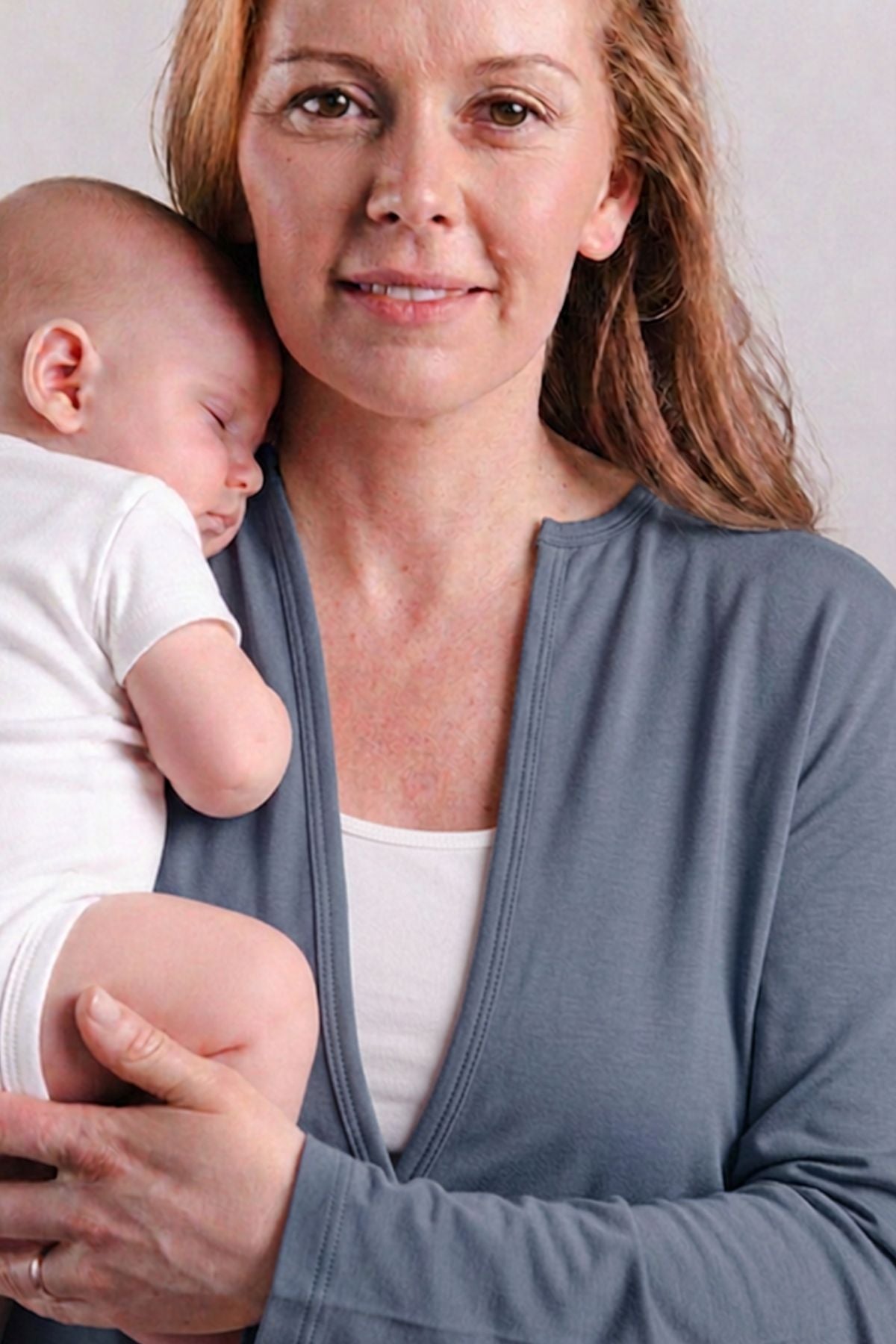 A woman wearing a slate blue baby wrap cardigan holds a sleeping newborn baby dressed in a white bodysuit against a plain light gray background.