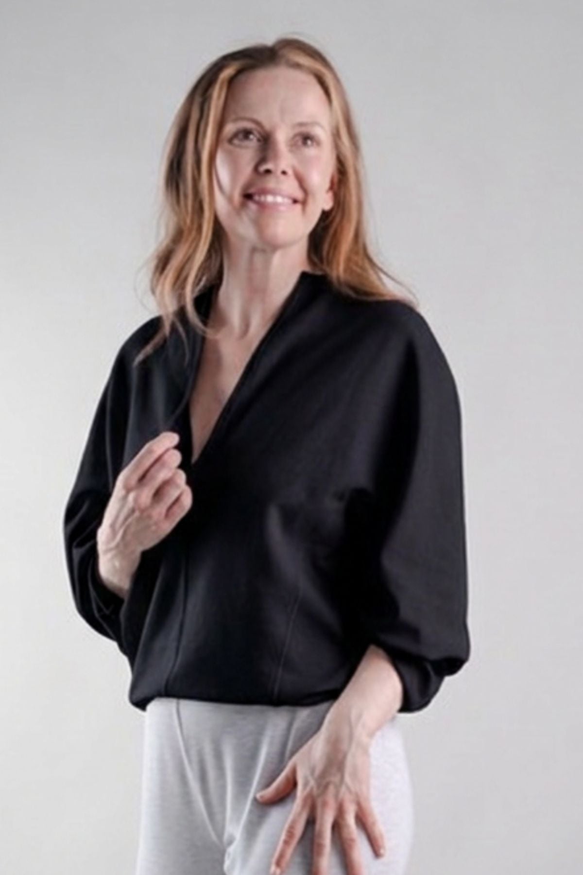 A woman with shoulder-length light brown hair is wearing a black wrap-style blouse with three-quarter sleeves and light gray sweatpants, standing against a neutral light gray studio background.