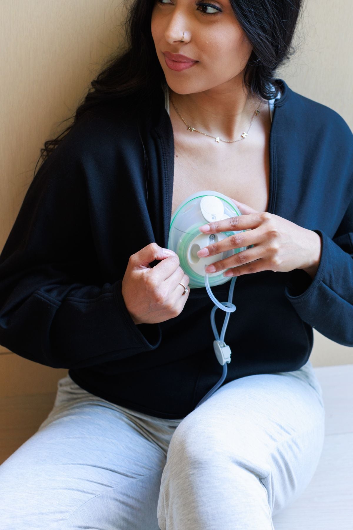 A woman wearing a black cardigan and light gray sweatpants holds a mint green and white breast pump cup connected to a clear tubing system.