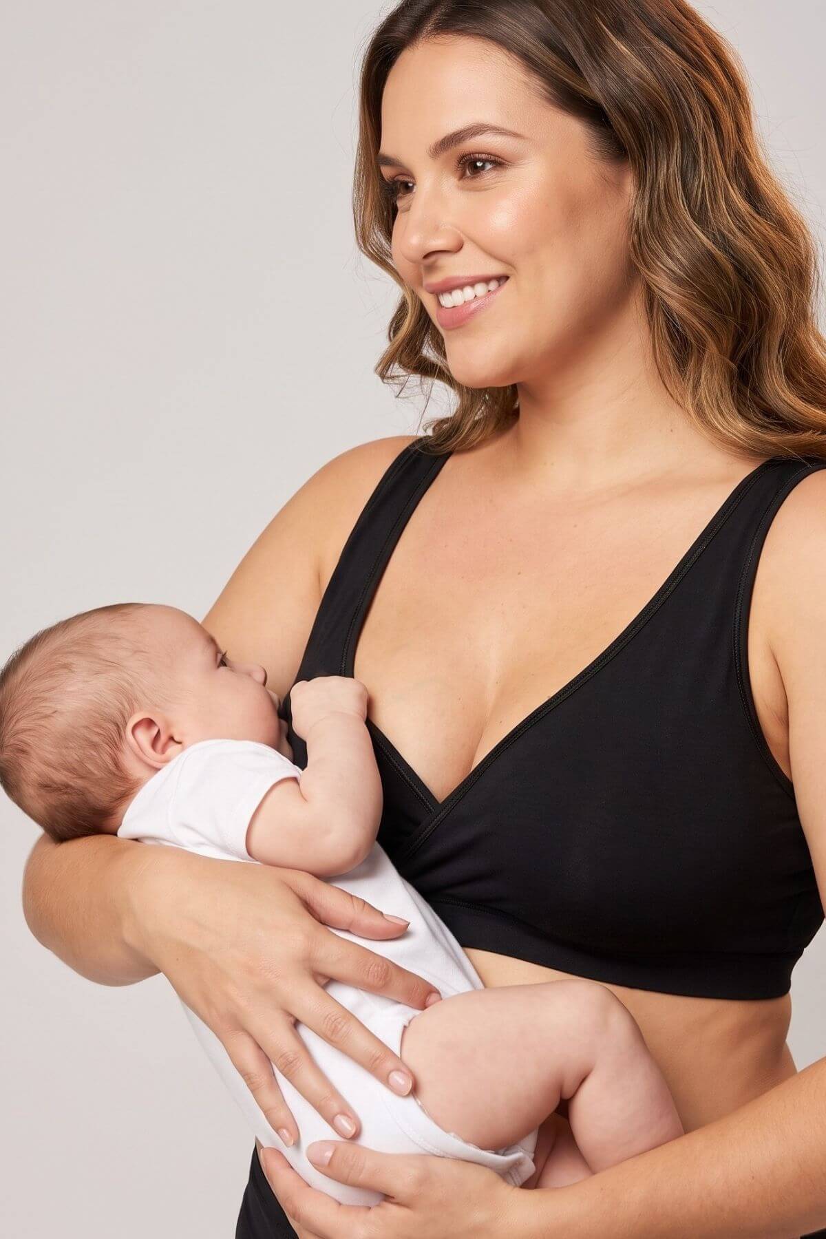 A woman wearing a black nursing bra holds a sleeping baby dressed in a white onesie against a plain light gray studio background.