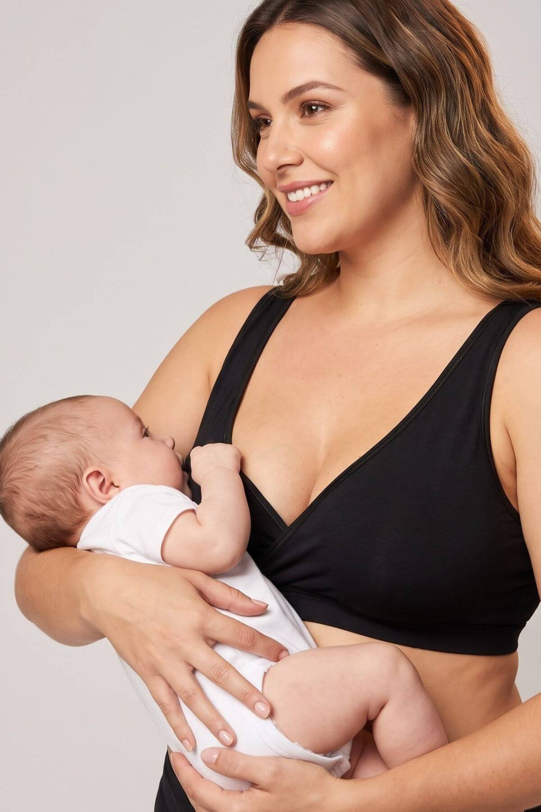 A woman wearing a black nursing bra holds a sleeping baby dressed in a white onesie against a plain light gray studio background.