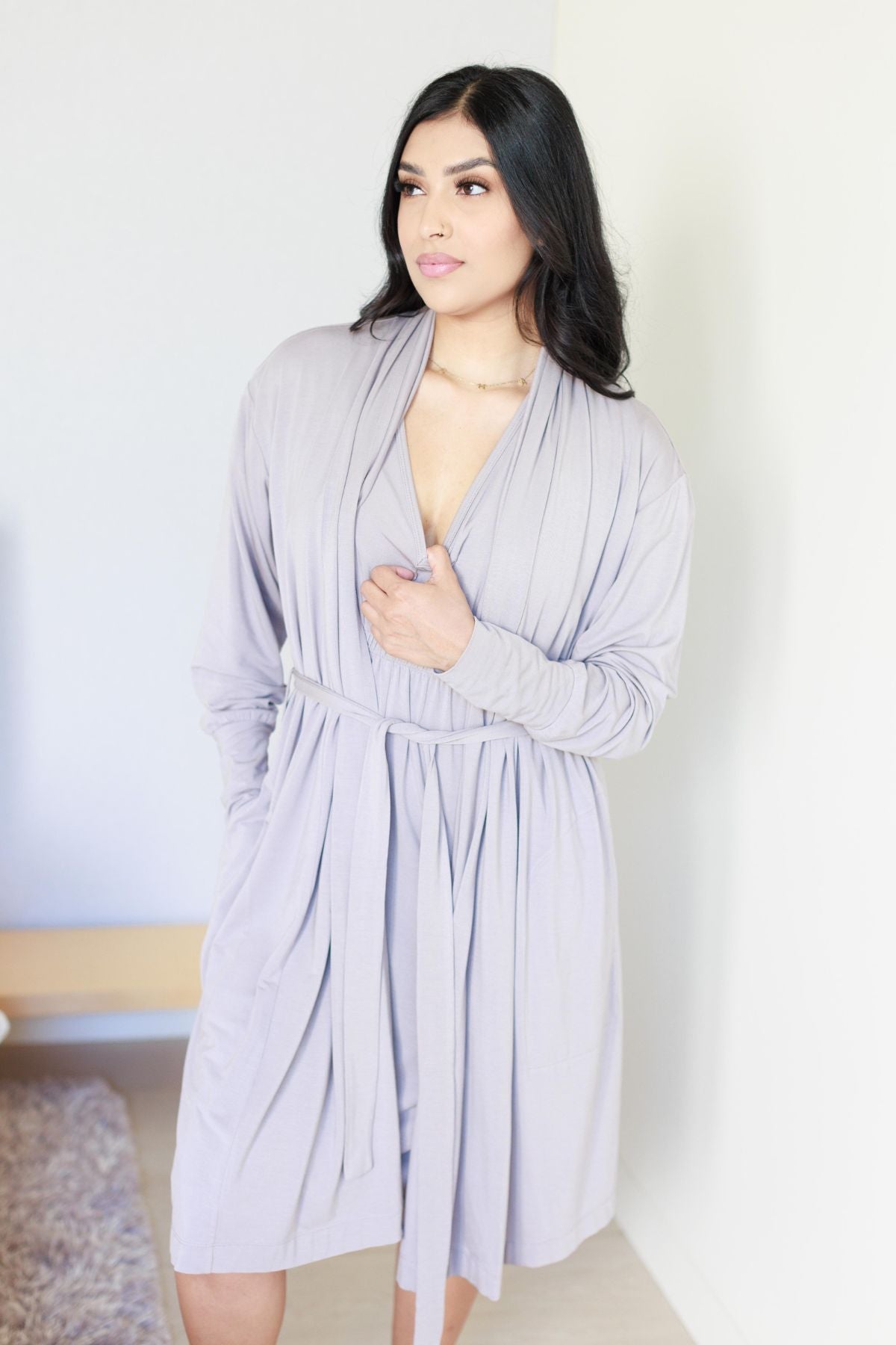 A woman modeling a light lavender pleated wrap dress with long sleeves, a tie waist, and side pockets in a brightly lit indoor setting..