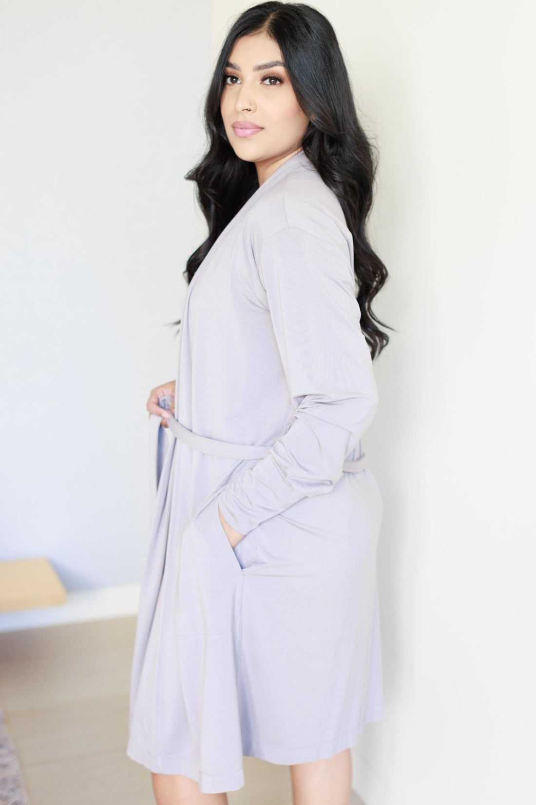 A woman modeling a light lavender long-sleeve wrap robe with a tie belt and side pockets, standing against a plain white wall..