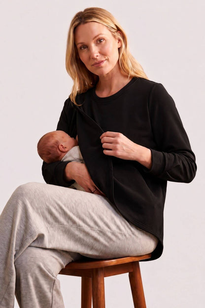A woman sitting on a wooden stool wearing a black long-sleeve maternity nursing top and light gray sweatpants, holding a baby against her chest. The background is a clean, neutral studio gray.