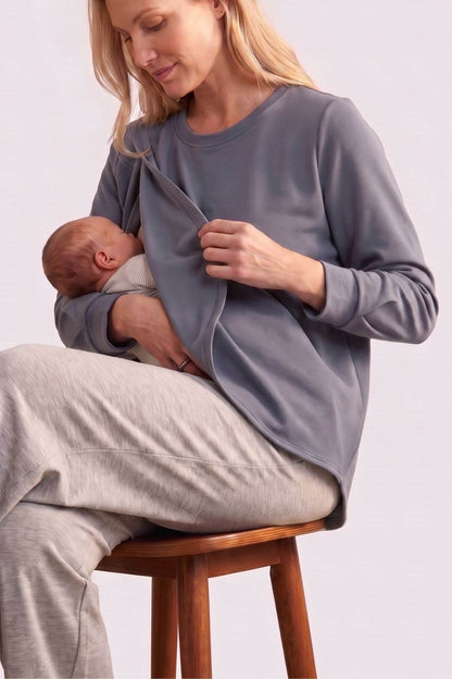 A woman sits on a wooden stool wearing a gray long-sleeve maternity nursing top and light gray pants, holding a baby against her chest. The background is a clean, neutral studio setting.