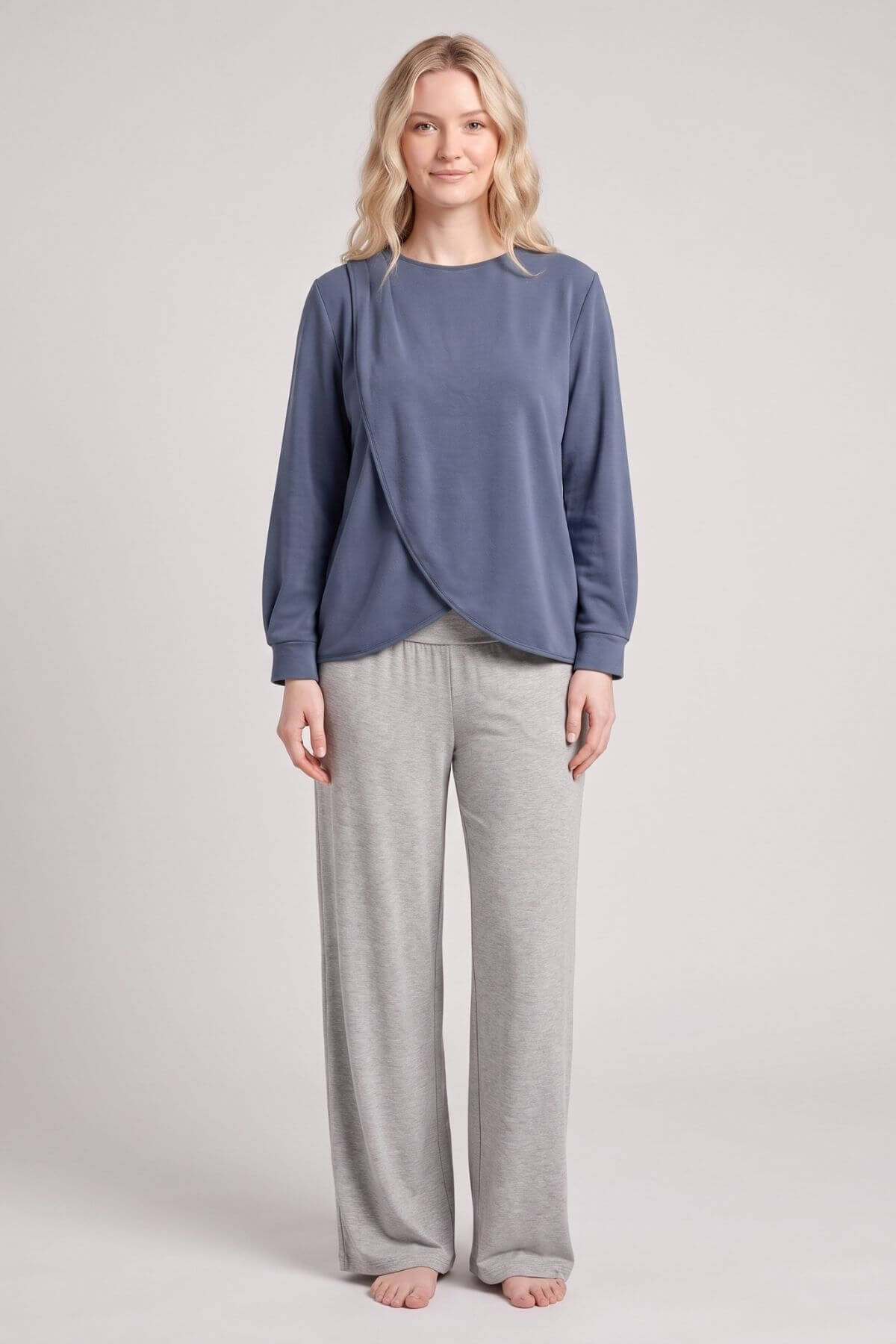 A woman stands against a light gray studio background wearing a slate blue long-sleeve wrap-style top and light gray wide-leg sweatpants.