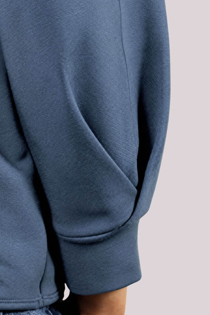 Close-up of a person wearing a slate blue sweatshirt, showing the sleeve and ribbed cuff against a light gray background.