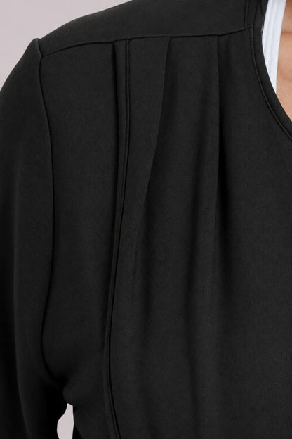 Close-up of a black long-sleeve top with a pleated front detail and a scoop neckline, worn over a white undershirt.