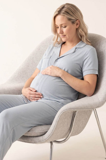 A pregnant woman wearing a light blue-gray short-sleeve button-front top and matching pants sits in a modern light gray upholstered chair against a plain off-white background.