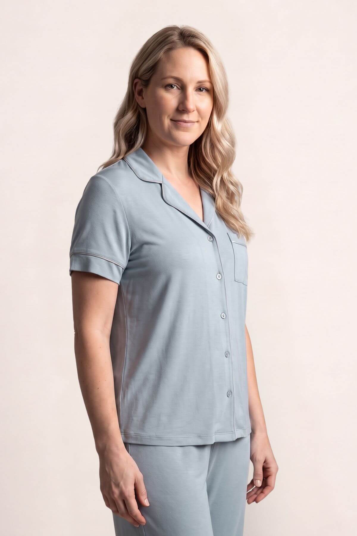 A woman modeling a light blue short-sleeve pajama top and matching pants against a plain beige studio background.