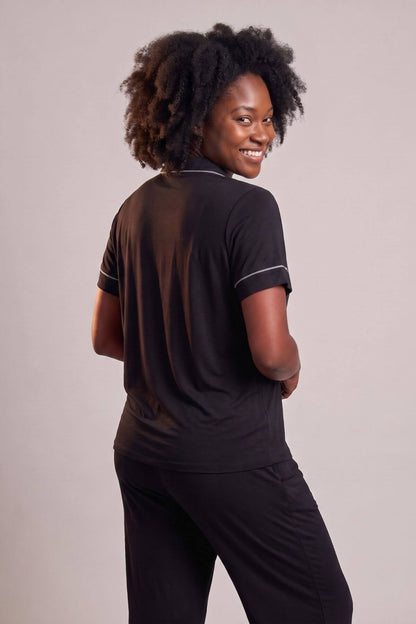 A woman with curly black hair is shown from the back wearing a black short-sleeve top with thin white piping details on the collar and sleeve cuffs, paired with black pants, against a solid light gray studio background.