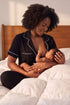A woman with dark curly hair sits on a bed wearing black pajamas with white piping and breastfeeding a baby. The background features a wooden headboard and white bedding.