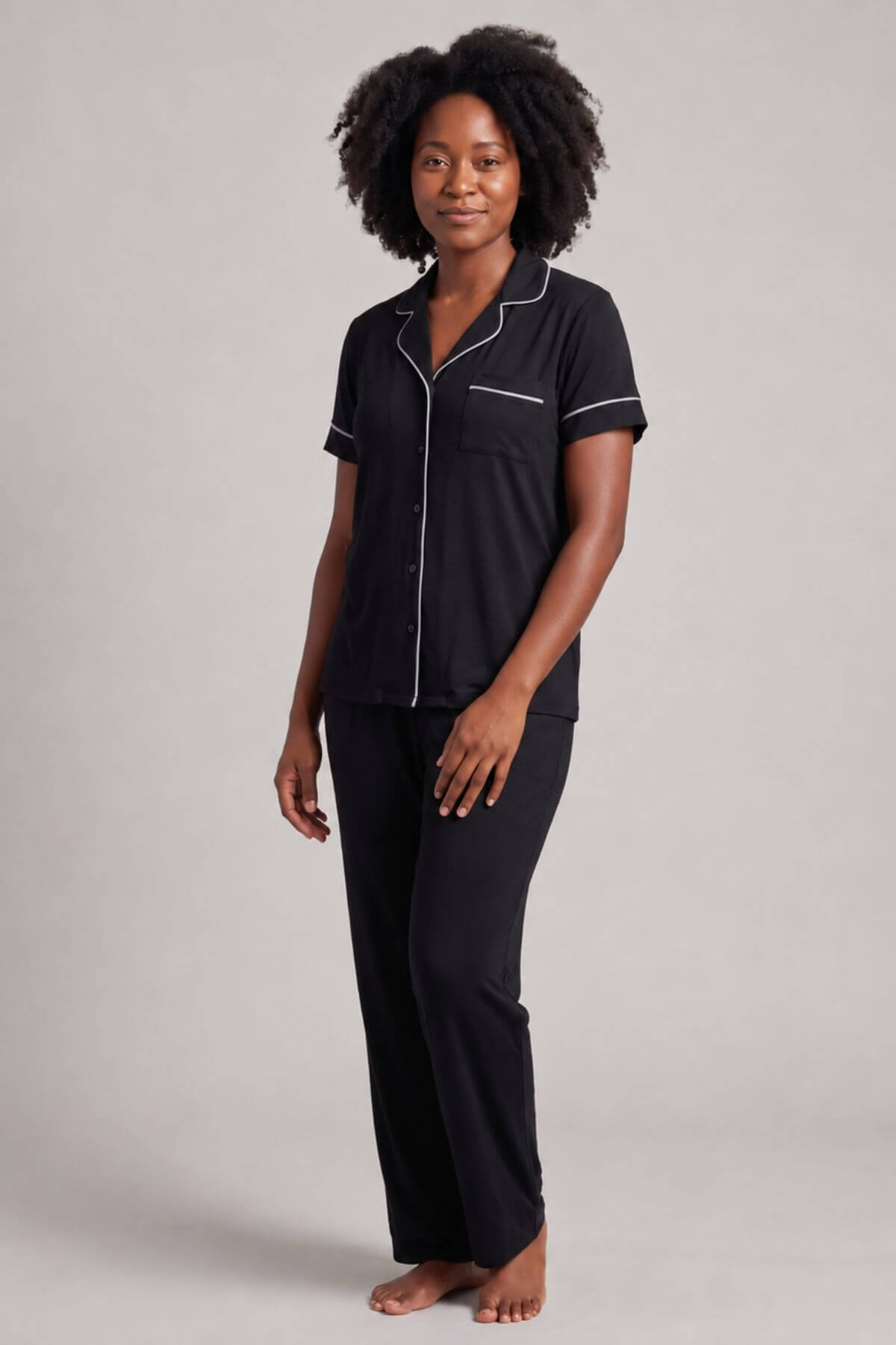 A woman modeling a black short-sleeve pajama shirt with white piping and matching black pajama pants against a light gray studio background.