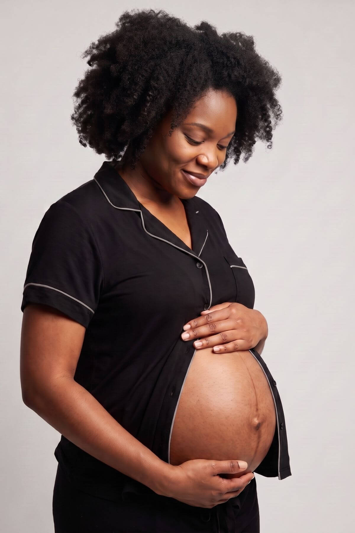 A pregnant woman with dark curly hair wearing a black short-sleeve pajama top with white piping, cradling her baby bump against a light gray studio background.
