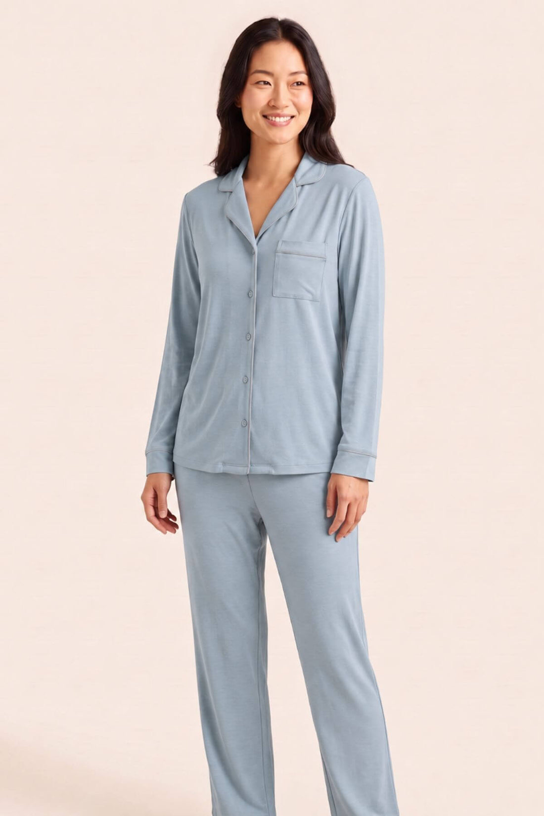 A woman modeling a light blue long-sleeve pajama set with a button-front shirt and matching pants against a solid pale beige background.