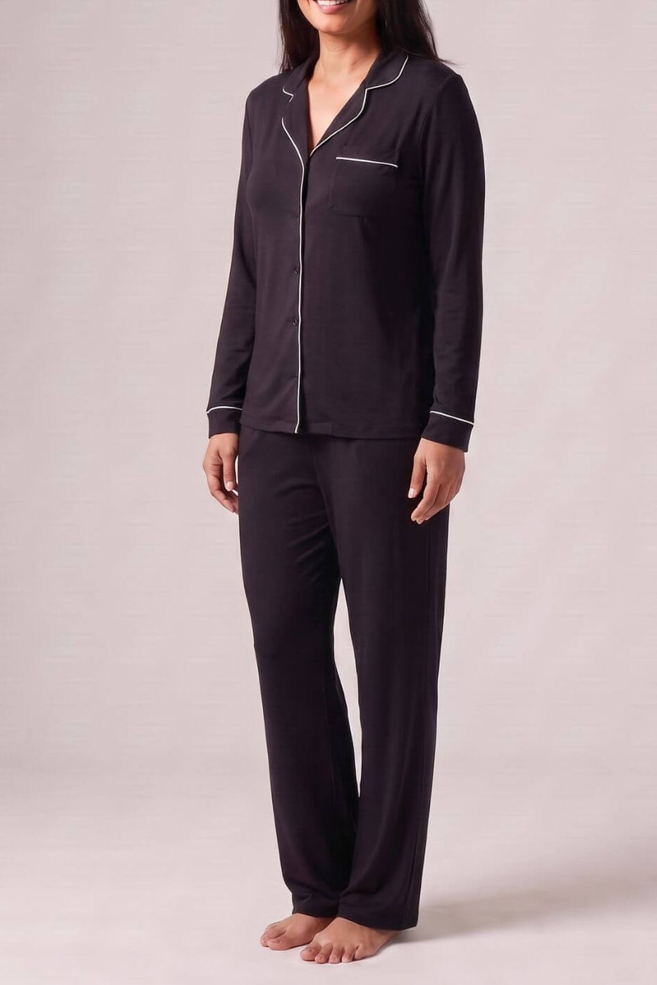 A woman modeling a dark purple long-sleeve pajama set with white piping details, standing against a neutral light gray studio background.