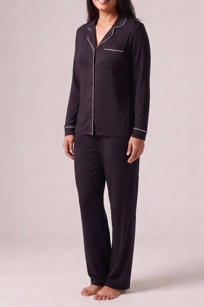 A woman modeling a dark purple long-sleeve pajama set with white piping details, standing against a neutral light gray studio background.