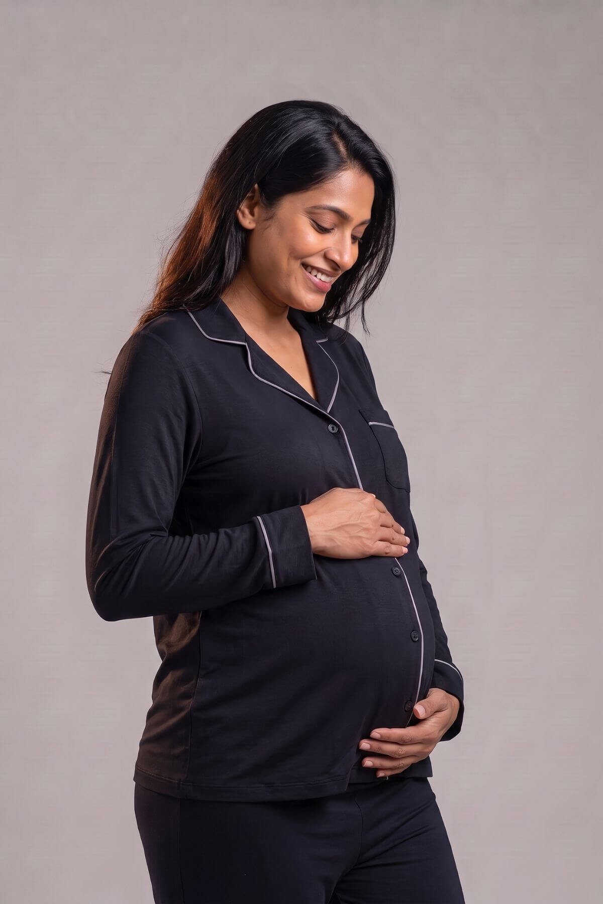 A pregnant woman wearing a black long-sleeve pajama set with white piping, standing against a neutral gray studio background.