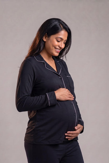 A pregnant woman wearing a black long-sleeve pajama set with white piping, standing against a neutral gray studio background.