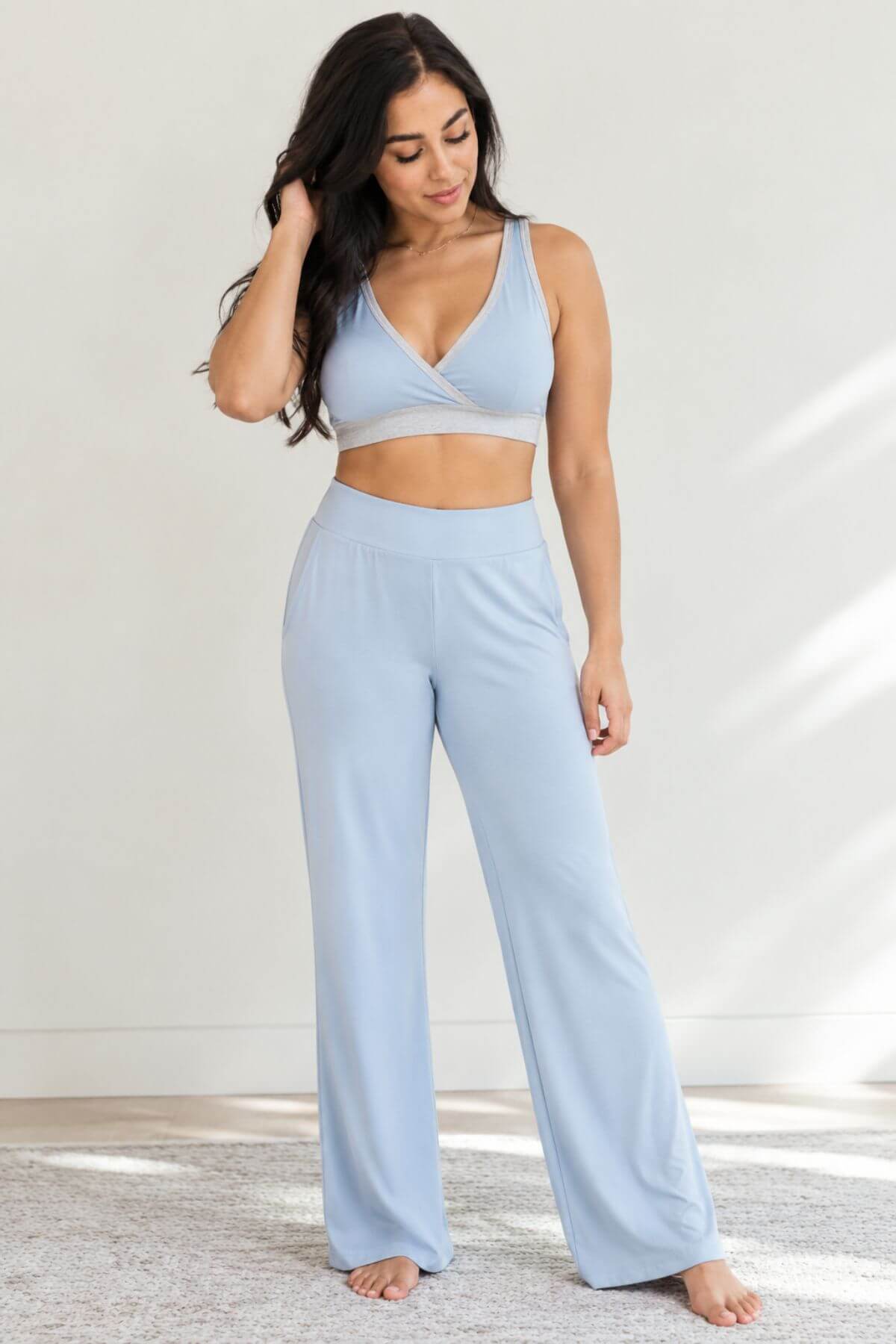 A woman modeling a light blue two-piece loungewear set consisting of a crossover sports bra and matching wide-leg pants, standing barefoot on a light rug against a white wall.